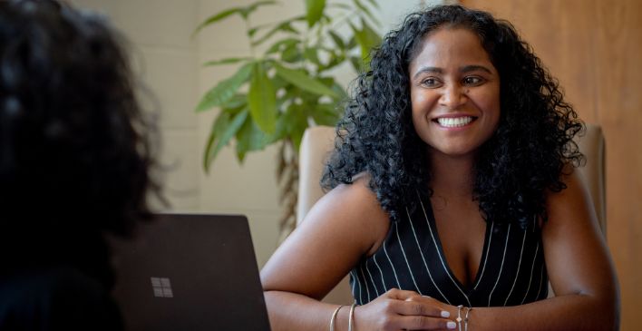 Screenshot of a smiling woman talking to another woman over her desk