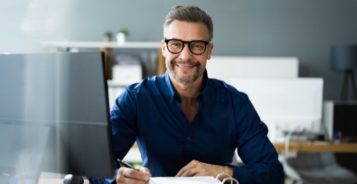 Man at his desk writing and smiling at the camera
